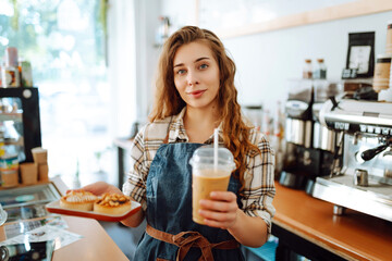 Curly-haired woman barista holding takeaway coffee and dessert. A young woman in an apron behind the bar gives out orders. Takeaway food and drink concept, small business.