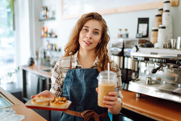 Curly-haired woman barista holding takeaway coffee and dessert. A young woman in an apron behind the bar gives out orders. Takeaway food and drink concept, small business.