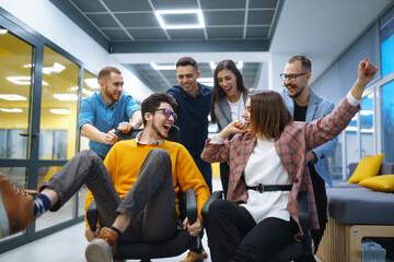 A group of young colleagues are having fun in the office corridor. Young business people ride on office chairs and laugh. Concept of teamwork, relaxation or fun. - obrazy, fototapety, plakaty