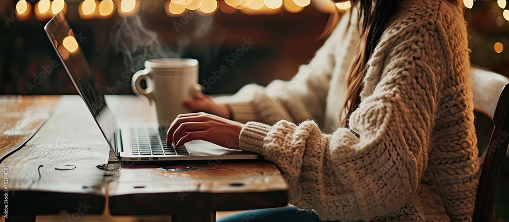 Poster female person sitting at wooden table and holding cup of hot drink while surfing the internet on not