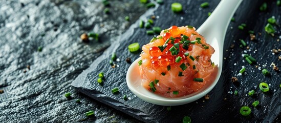 A close up on a taste of gourmet salmon tartare for catering with chives and caviar decorating on an elegant white spoon and presented on a black slate. with copy space image