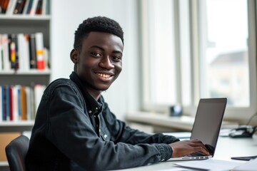 Teenage african man sitting in white office with laptop, smile, look at camera he is a student studying online with laptop at home, university student studying online, online web education concept - obrazy, fototapety, plakaty