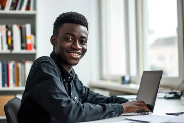 Tuinposter Teenage african man sitting in white office with laptop, smile, look at camera he is a student studying online with laptop at home, university student studying online, online web education concept © Attasit