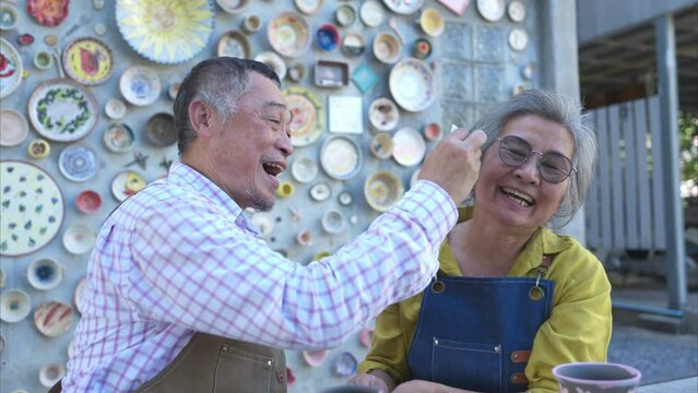 In the pottery workshop, an Asian retired couple is engaged in pottery making and clay painting activities, Have fun by smearing paint on each other's faces.
