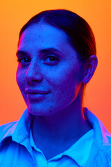 Close up portrait of young attractive woman, student with freckles looking at camera against orange background in neon light. Concept of beauty, youth, human emotions, self-expression, lifestyle. Ad