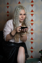 Platinum blonde girl, 25-30 years old, wearing a black leather skirt and pink sweater, sitting, holding a black cup of hot coffee. In a 70s style restaurant.