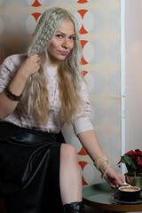 Platinum blonde girl, 25-30 years old, wearing a black leather skirt and pink sweater, sitting, holding a black cup of hot coffee. In a 70s style restaurant.