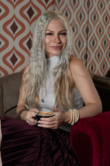 Platinum blonde girl, 25 to 30 years old, wearing a white tank top and a red skirt. With a cup of coffee in her hand, sitting in an antique armchair. Makeup In a 70s style restaurant.