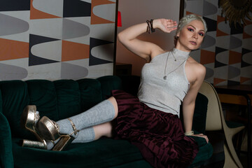 Platinum blonde girl, 25 to 30 years old, wearing a white tank top and a red skirt, gray stockings, lying on an antique green armchair. Makeup In a 70s style restaurant.