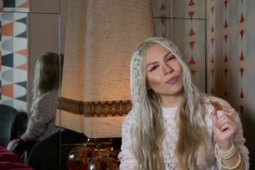 Platinum blonde girl, 25-30 years old, dressed in a pink sweater, sitting, eating a cookie, with an antique lamp behind her. Reflected in a mirror. In a 70s style restaurant.