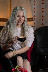 Platinum blonde girl, 25-30 years old, wearing a pink sweater, sitting with a black cup of hot coffee. In a 70s style restaurant.