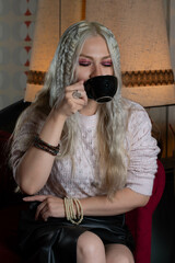 Platinum blonde girl, 25 to 30 years old, sitting in a red armchair, wearing a pink sweater and a black skirt, drinking a cup of coffee. Makeup In a 70s style restaurant.