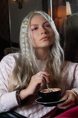 Platinum blonde girl, 25-30 years old, wearing a pink sweater, sitting with a black cup of hot coffee. In a 70s style restaurant.