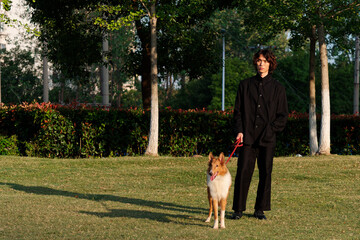 Portrait of a handsome Chinese young man in black with his rough collie dog on green grass field in sunny day, male fashion, cool Asian young man lifestyle, harmony man and pet.
