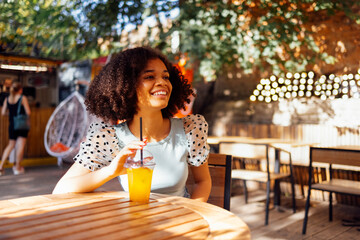 Happy african american teenage girl drinking lemonade or cocktail in an open air cafe. Smiling darkskin female teenager sits at table on the summer terrace. - obrazy, fototapety, plakaty