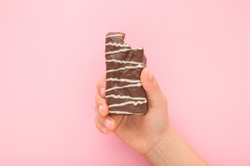 Baby girl hand holding and showing bitten bar with dark brown chocolate glaze and white stripes on light pink table background. Pastel color. Sweet snack. Closeup. Top down view. - obrazy, fototapety, plakaty