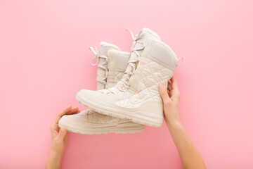 Young adult woman hands holding new beige warm winter boots with shoelace on light pink table background. Pastel color. Female footwear. Closeup. Point of view shot. Top down view.