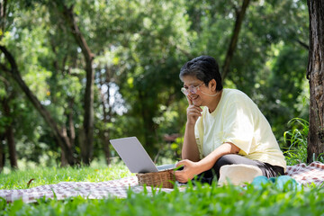 Happy Asian senior woman using laptop for working online outside office. Elderly retirement using laptop for learning new skill and freelance working in park, senior lifestyle with technology