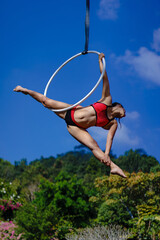 Beautiful Asian girl performing show aerial hoop or aerial ring in various positions and spinning stunts on the blue sky around the natural fresh and the waterfall, outdoors.