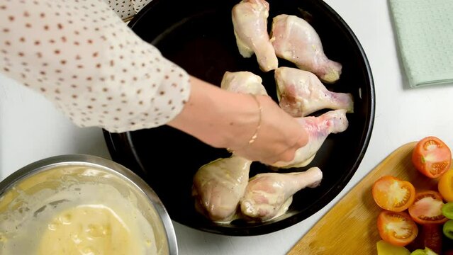 Woman putting chicken drum sticks in sause on baking sheet, tray in kitchen, top view. Cooking meat dish with vegetables. Preparing food, meal from healthy eco products. Culinary, cuisine, recipe.