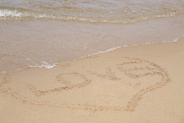 Written LOVE surrounded by a heart shape on a fine sand background. Handwritten on the wet beach sand with a small wave lapping.
