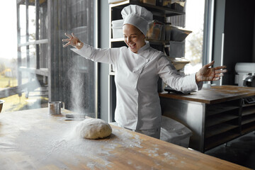 Excited enthusiastic young woman baker throwing flour on dough