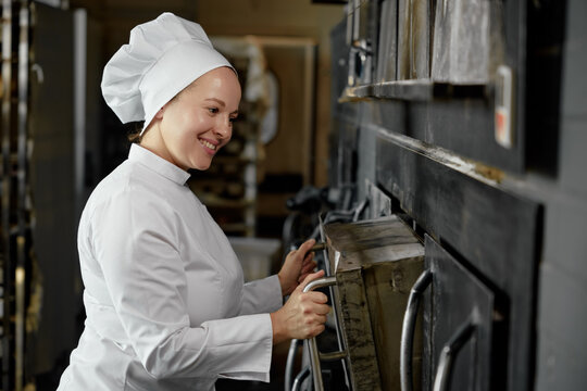 Woman baker in uniform opening oven stove at bakery kitchen