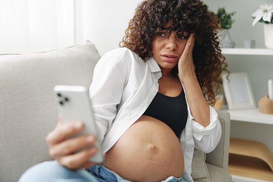 Pregnant woman blogger sits on the couch at home and takes pictures of herself on the phone, selfie and video call, consultation with the doctor online, pregnancy management, freelance work