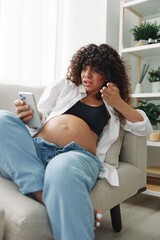 Pregnant woman blogger sits on the couch at home and takes pictures of herself on the phone, selfie and video call, consultation with the doctor online, pregnancy management, freelance work