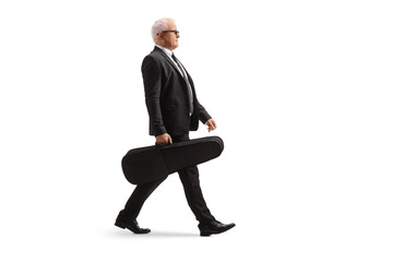Full length profile shot of a man in a black suit walking and carrying a violin in a case