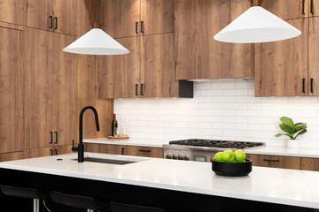A kitchen detail with wood cabinets, a black faucet, subway tile backsplash, and gold light fixtures hanging above the black island and marble countertops.