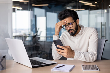 Upset and worried young Indian man sitting in the office at the desk, holding his head and looking frustrated at the phone screen