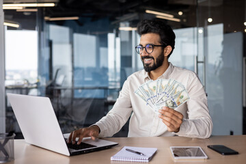 Indian smiling young man businessman working in the office on a laptop, sitting at the table and...