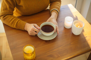 Woman holding cup of hot tea while sitting at table with with burning candles at home