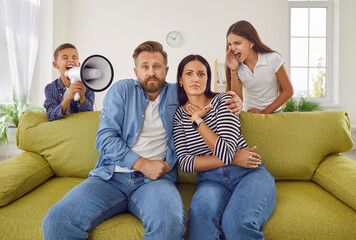 Children screaming at parents. Two naughty kids making lots of noise. Unruly brother and sister with megaphone screaming at mum and dad sitting on sofa in living room with scared face expressions - obrazy, fototapety, plakaty