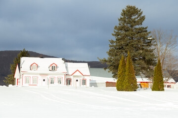 Beautiful patrimonial double wooden white house with shingled pitched roof and wooden barns seen during a cloudy day after a fresh snowfall, Quebec City, Quebec, Canada