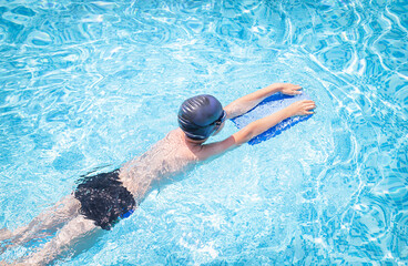 Active child (boy) in cap, sport goggles doing water sport with swim board in the swimming pool....