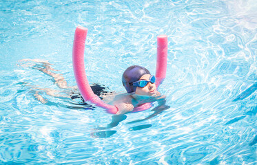 Active child (boy) in cap, sport goggles doing water sport with swim noodle in the swimming pool....