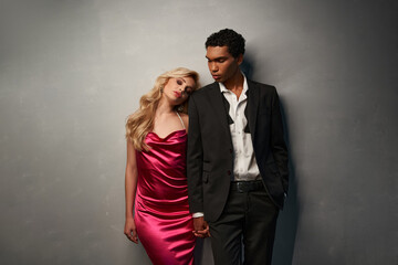 alluring beautiful diverse couple in chic outfits holding hands and posing together on gray backdrop
