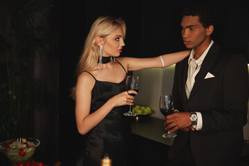 appealing sensual diverse couple in elegant attires holding wine glasses and looking at each other