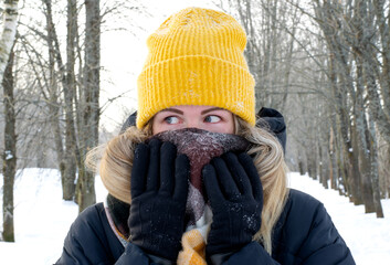 Young woman in yellow knitted cap and black puffer jacket is hiding her nose from cold in a scarf...