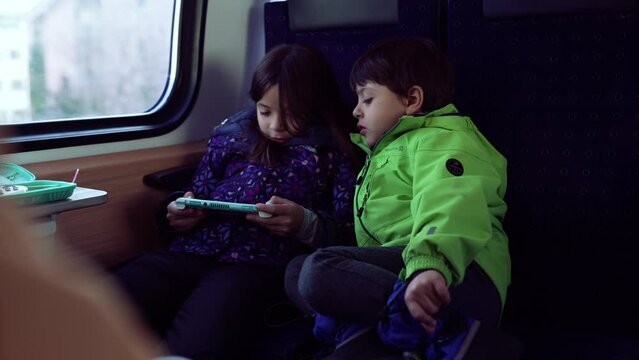 Passenger Small girl playing game with handheld console while younger brother stares at her screen play, kids engaged with technology while on the go inside train