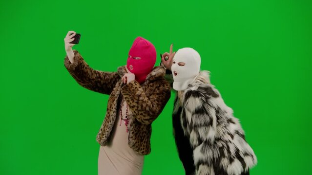 Two women in white and pink balaclavas taking selfies using smartphone. Freak women in fur coats on green background studio. Fashion trend concept, feminist trend in fashion.