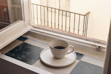 Cup of coffee on vintage in ceramic mug checkered tile windowsill. Blurred beige wall with balcony...