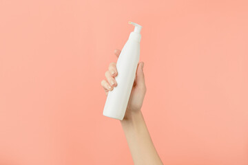 A womans hand holding a white cosmetic bottle on a peach background. Beauty treatment concept
