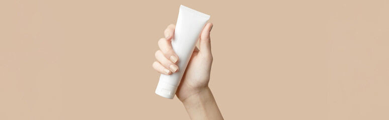A womans hand holding a white cosmetic tube on a beige background. Care products