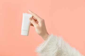 A womans hand in a sweater holds a tube of cream on a peach background. Self care beauty treatment