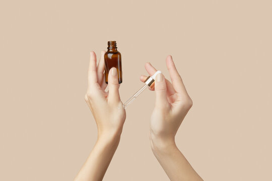 A womans hands holding a brown bottle of serum on a beige background. Body care