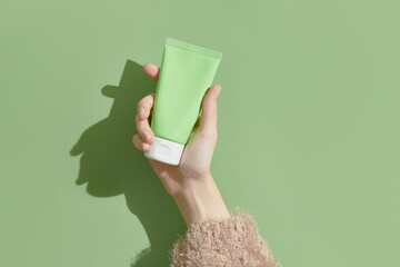 A womans hand in a sweater holds a tube of cream on a green background. Self care beauty treatment