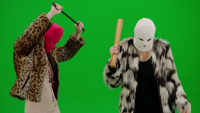 Two women in white and pink balaclavas dancing funny with a bat and a crowbar. Freak women in fur coats on green studio background. Fashion trend concept, feminist trend in fashion.